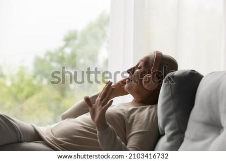 Side view happy old mature retired woman listening popular music in headphones, relaxing alone on cozy sofa, enjoying peaceful carefree weekend time alone at home, stress free leisure pastime. Royalty-Free Stock Photo #2103416732