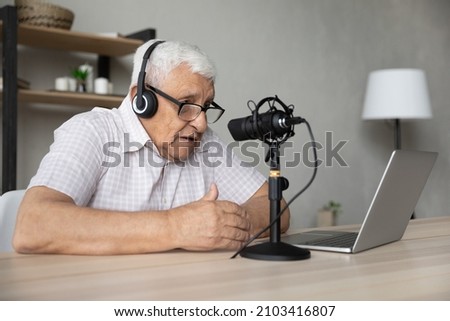 Focused mature elderly retired man in eyeglasses wearing wireless headphones looking at computer screen, talking in professional stand microphone, voice acting or recording vlog alone at home.