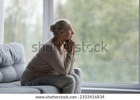 Pensive old mature woman looking in distance out of window, sitting on couch alone at home, suffering from negative thoughts or psychological problems, retirement lifestyle, ageing process concept. Royalty-Free Stock Photo #2103416834