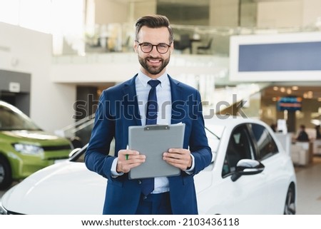 Closeup shot of successful confident smiling caucasian male shop assistant holding clipboard in formal clothes looking at camera at automobile car dealer shop Royalty-Free Stock Photo #2103436118