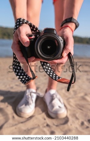 Man holding camera in front of photographer. Sand beach and river in background. Sunny day.