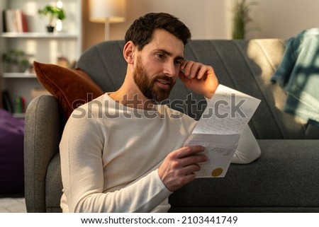 Caucasian man feeling happy while reading his letter from relatives and sitting at the floor near the sofa. Stock photo