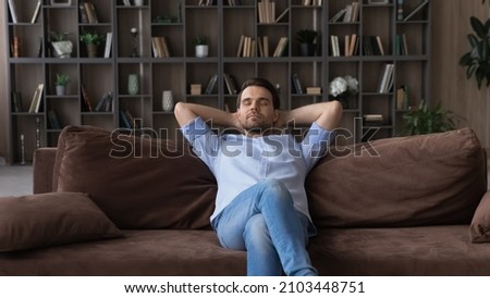 Serene man with closed eyes sitting relaxing on comfortable couch at home alone, enjoying lazy weekend leisure time, daydreaming or taking day nap, dreaming of good future, breathing fresh air Royalty-Free Stock Photo #2103448751