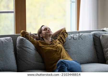 Calm serene sleepy young woman relaxing on comfortable sofa at home, leaning om back, taking deep breath of fresh cool air, enjoying relaxation, leisure time, meditating for stress relief Royalty-Free Stock Photo #2103448814