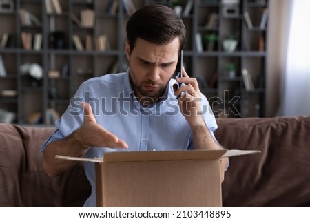 Dissatisfied unhappy man talking on phone with customer support service, complaining, sitting on couch at home, frustrated angry customer arguing with shipping company, bad delivery service concept Royalty-Free Stock Photo #2103448895