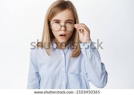 Portrait of blond girl, little kid in glasses squinting, has difficulties reading without eyewear, standing over white background