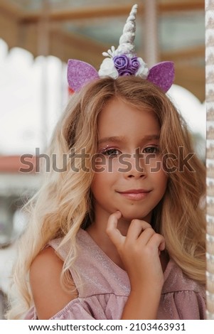 Blond hair with unicorn headband beautiful little girl in pink dress sits on carousel in the park with cotton candy 