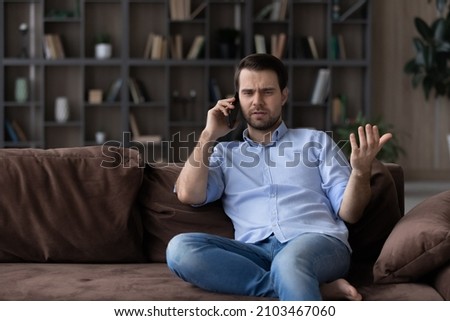 Angry dissatisfied man talking on phone, arguing or solving problem, sitting on couch at home, irritated unhappy client having unpleasant conversation with customer support service, complaining Royalty-Free Stock Photo #2103467060
