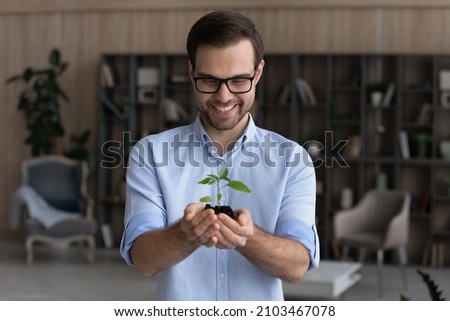 Smiling businessman wearing glasses holding small green plant sprout with soil, standing, happy entrepreneur employee with growing tree, startup project, profit, investment and growth concept Royalty-Free Stock Photo #2103467078