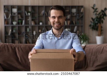 Head shot portrait smiling man holding cardboard box, giving or receiving parcel, sitting on couch at home, happy satisfied customer looking at camera, good delivery service and fast shipping concept Royalty-Free Stock Photo #2103467087