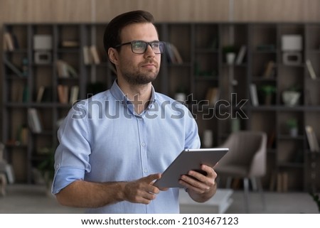 Dreamy confident man wearing glasses looking in distance, holding tablet in hands, distracted from online chatting, thoughtful pensive man visualizing future, planning workday, standing at home Royalty-Free Stock Photo #2103467123