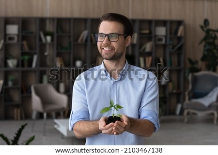 Dreamy smiling man in glasses holding green plant with soil, growing small tree, environment concept, happy pensive businessman entrepreneur with sprout in hands, investment and growth