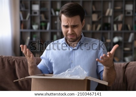 Head shot unhappy dissatisfied man opening parcel at home, sitting on couch with cardboard box, angry displeased customer confused by wrong or damaged order, bad delivery shipping service concept Royalty-Free Stock Photo #2103467141
