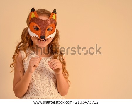 A child in a masquerade carnival mask of a fox, hand-made from glitter foamiran. Little cute cheerful comical girl in the role of a fox. The child is preparing for the carnival, holidays. 