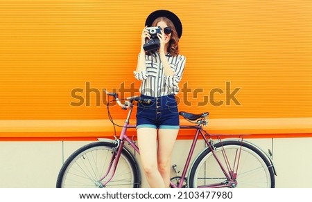Summer colorful portrait of happy smiling young woman photographer taking a picture by film camera with bicycle in the city on orange background