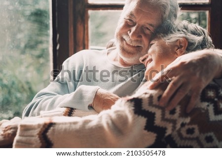 Old senior couple in love hug and embrace with romance together at home with outside view in windows background - happy mature retired people lifestyle enjoying caring each other and smiling Royalty-Free Stock Photo #2103505478