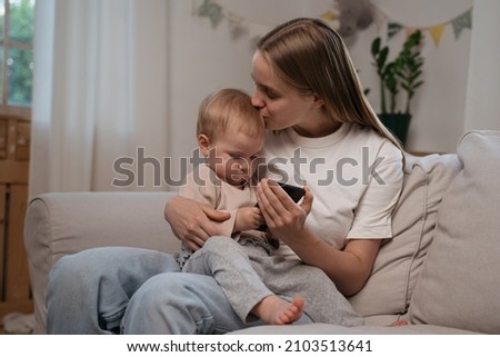 Young mother hugging and kissing her cute baby child sitting on sofa. Authentic portrait of happy family using mobile phone watching online cartoon at home. Love and care concept	
