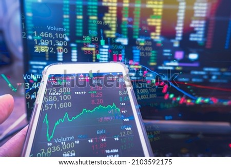 Businessman holding phone checking stock market data on computer screen background.
