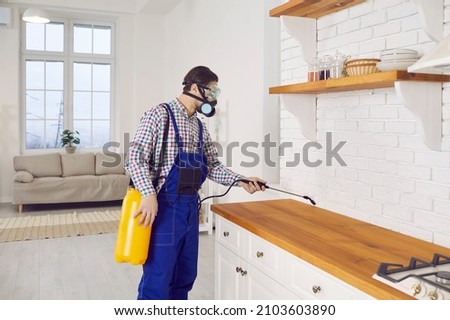 Pest control guy spraying rodenticide, or termite and cockroach insecticide inside house. Worker in mask and blue overall uniform standing in kitchen and spraying poison over countertop and cabinets