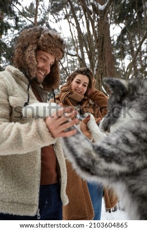 Happy young couple in winterwear playing with purebred siberian husky and having fun in snowbound pine forest Royalty-Free Stock Photo #2103604865