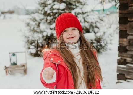 A cute girl in a sweater and a red hat with sparklers in winter on the street by the Christmas tree in the lights laughs and has fun. Image with selective focus