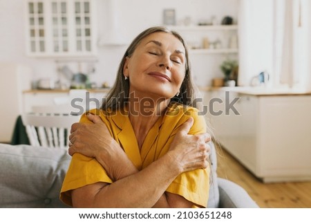 Horizontal indoor portrait of happy elderly woman hugging herself, giving support, showing self-respect and love, sitting on couch with closed eyes in yellow shirt. Human emotions, body language Royalty-Free Stock Photo #2103651248