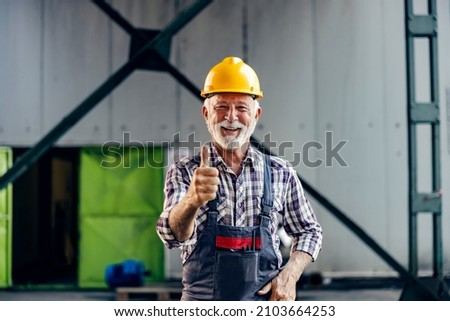 Good business in the factory. A satiated smiling senior worker with a helmet on his head is standing in the factory and showing thumbs up. Royalty-Free Stock Photo #2103664253