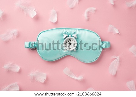 Top view photo of small white alarm clock on light blue silk sleeping mask and light pink feathers on isolated pastel pink background
