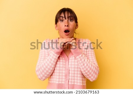 Young Argentinian woman isolated on yellow background praying for luck, amazed and opening mouth looking to front.