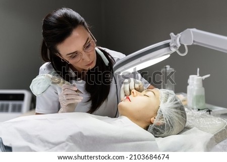 Professional permanent makeup artist and her client during lip blushing procedure Royalty-Free Stock Photo #2103684674