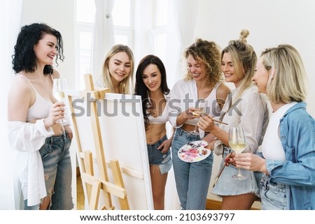 Group of young beautiful women paint on canvas and drinking white wine during party at home