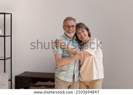 Portrait of affectionate loving old woman cuddling shoulders of sincere mature husband, looking at camera posing in modern living room, feeling excited in own dwelling, retirement lifestyle concept. Royalty-Free Stock Photo #2103694847
