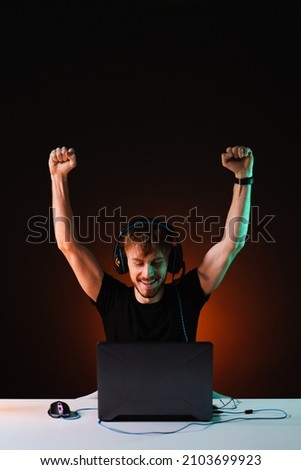 Joyful young gamer rejoices in winning a video game with a smile on his face and raised hands, wears casual clothes and a headset. Streamer winner plays at home on the computer on dark background.
