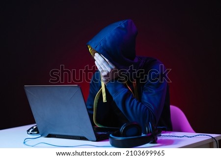 Hacker with a laptop sits at the table, dark background and neon light behind, head covered with a hood.