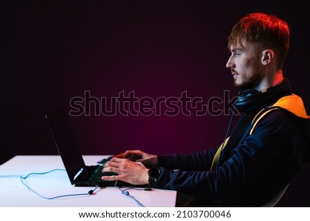 Portrait of Software Developer. Hacker. Gamer Wearing Headset Sitting at His Desk and Working. Playing on Laptop. In the Background Dark High Tech Environment with red neon lights.