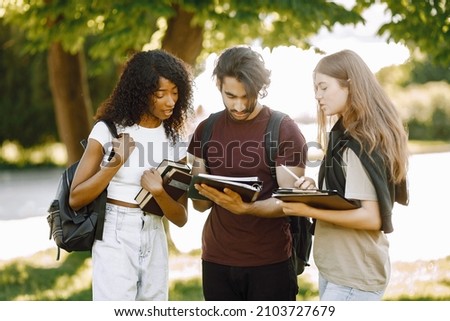 Three international students standing in a park and holding a books