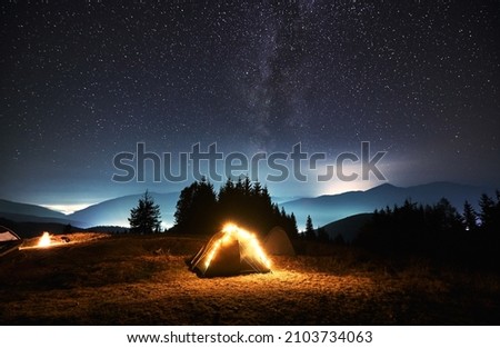 Tourist tent lit by bright yellow bulbs on green night lawn next to another tent. Camping on starry night time. Forest and silhouettes of mountain hills on the background.