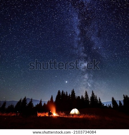 Male traveler looking at campfire while sitting near camp tent under beautiful night sky with stars and Milky way. Concept of travelling, hiking and night camping.