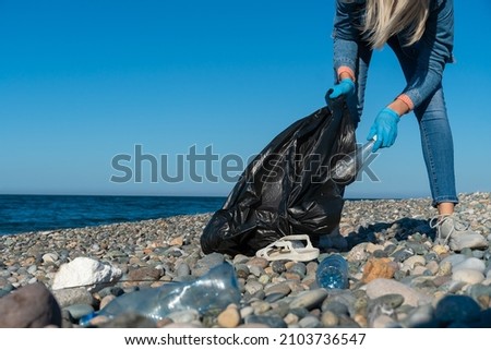 Young woman collects plastic, rubber garbage in a black garbage bag on a pebble beach by the sea. Empty used dirty plastic bottles. Environmental pollution on the Black Sea coast