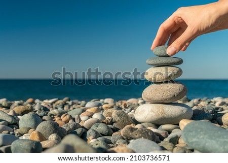 Close-up of a woman's hand putting the last stone on top of pebbles on the seashore on a sunny day, copy space. Concept of balance, harmony