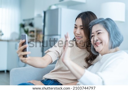 Asian lovely family, young daughter use phone selfie with older mother. Beautiful girl and senior elderly woman sit on sofa, enjoy video call by internet online talking together in living room at home