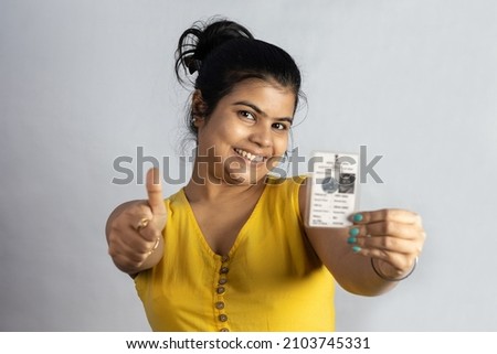 An Indian young woman smiling and showing thumbs up to voter card in hand on white background