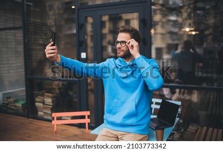 Caucaisan man in classic spectacles clicking selfie images during leisure time using mobile application outdoors, millennial hipster guy looking at front smartphone camera during daytime in city