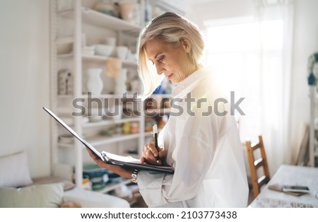Focused mature european female entrepreneur writing something in clipboard. Small business and entrepreneurship. Modern successful woman. Home art studio with pottery on shelves at sunny day Royalty-Free Stock Photo #2103773438