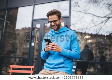 Happy hipster guy in classic spectacles for provide vision correction using cellphone gadget for checking received email message, smiling Caucasian male blogger browsing website in social media Royalty-Free Stock Photo #2103778655