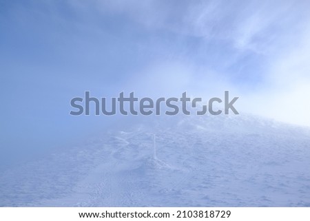 Frozen pole marking trail in mountains by path on misty and sunny winter day in Babia Gora massif. Silhouettes of people climbing to Babia Gora peak in background. Diablak, Beskid Zywiecki, Poland
