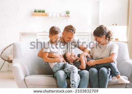 Smiling father sitting on sofa with happy wife and little children and brothers. Excited parents look at children. Happy family with two sons playing at home. The family is sitting on the couch