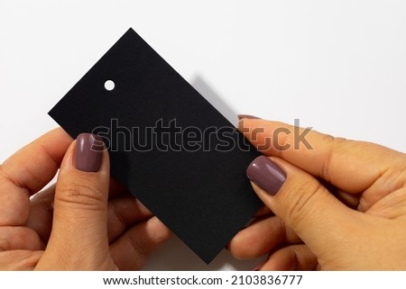 Two hands with cardboard black rectangular blank tag with little hole in upper part for clothes in center of white background. Tag mock up. Copy space.