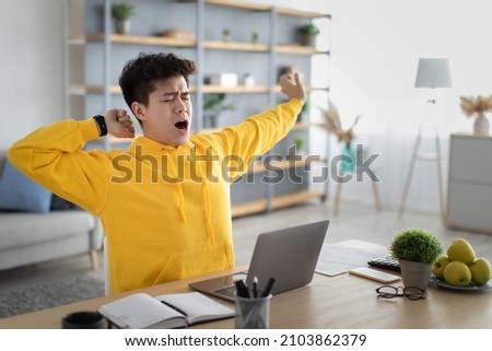 Tired asian male student yawning sitting at desk using laptop. Bored guy is exhausted from getting ready for test or writing coursework, feeling sleepy, stretching arms. Lack of sleep and tiredness Royalty-Free Stock Photo #2103862379