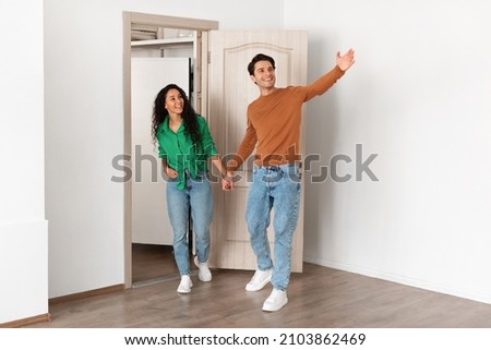 Look At This. Portrait of excited guy inviting lady to enter home, welcoming to living room, happy smiling man holding woman's hand showing new modern apartment, walking in flat together Royalty-Free Stock Photo #2103862469